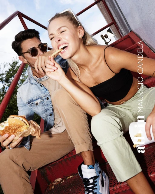 Two people, a man and a woman, are sitting on red stairs eating a burger and fries and smiling against a sky background.