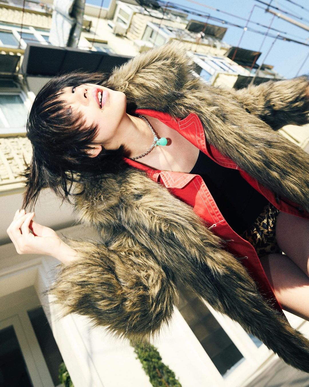 Stylish woman poses outdoors wearing a furry coat and red jacket, looking up towards the sky with a building in the background.