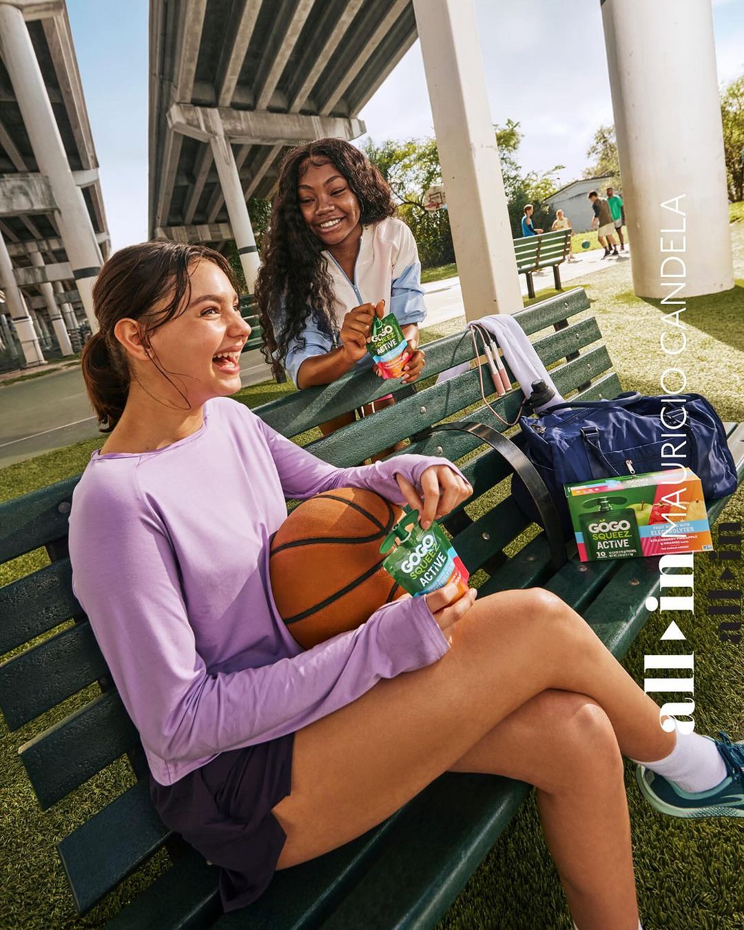 Two smiling young women on outdoor basketball court, one with a basketball, both holding GoGo squeeZ ACTIVE fruit snack pouch.