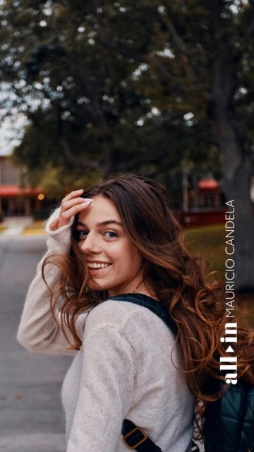 Smiling woman with long brown hair, wearing a sweater and backpack, stands on a tree-lined campus walkway while looking back.
