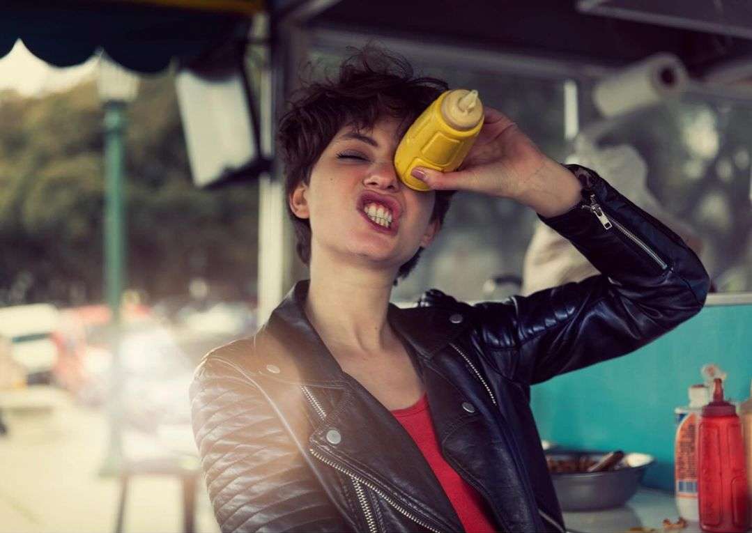 A young adult woman in a black leather jacket grimaces playfully with a yellow squeeze bottle near her eye outdoors at a food stand.