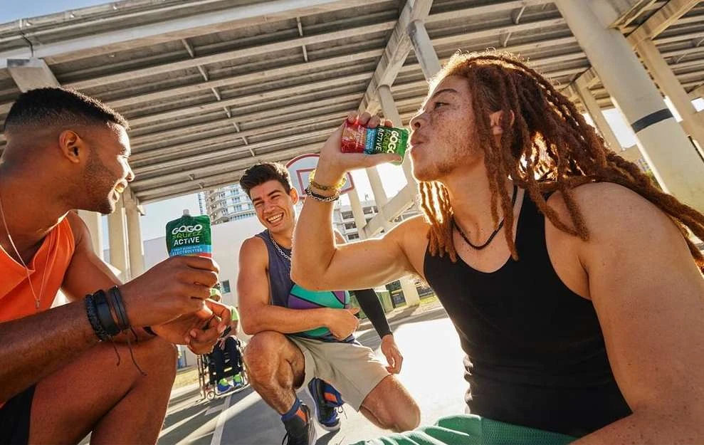 Three young adult men smile outdoors on a basketball court, one drinking a GoGo squeeZ ACTIVE pouch drink in a sunny setting.