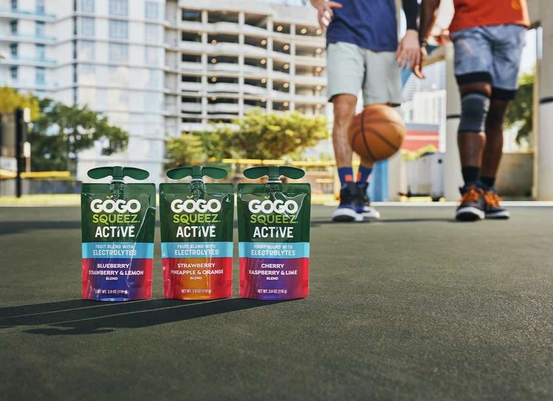Three GoGo Squeez Active fruit pouches are foregrounded, with two men playing basketball in the background on an outdoor court.