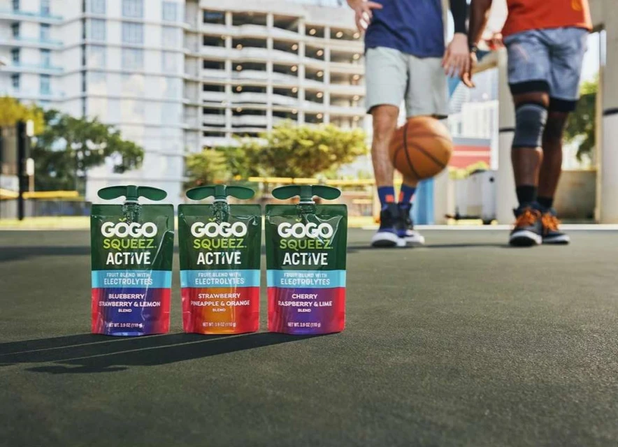 Three GoGo Squeez Active fruit pouches are foregrounded, with two men playing basketball in the background on an outdoor court.