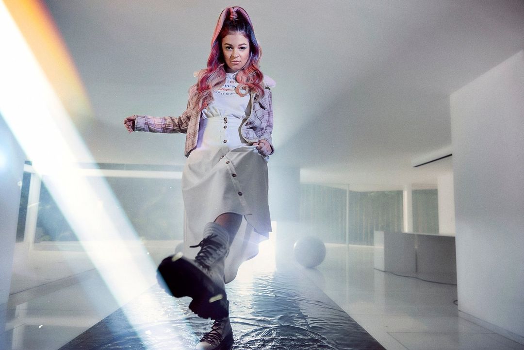 Woman with dyed hair, wearing a white dress and boots, walking in a modern interior with a shiny floor and white walls.