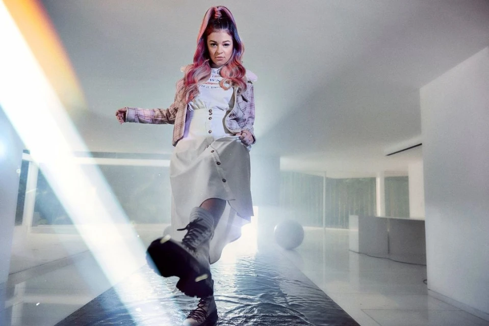 Woman with dyed hair, wearing a white dress and boots, walking in a modern interior with a shiny floor and white walls.