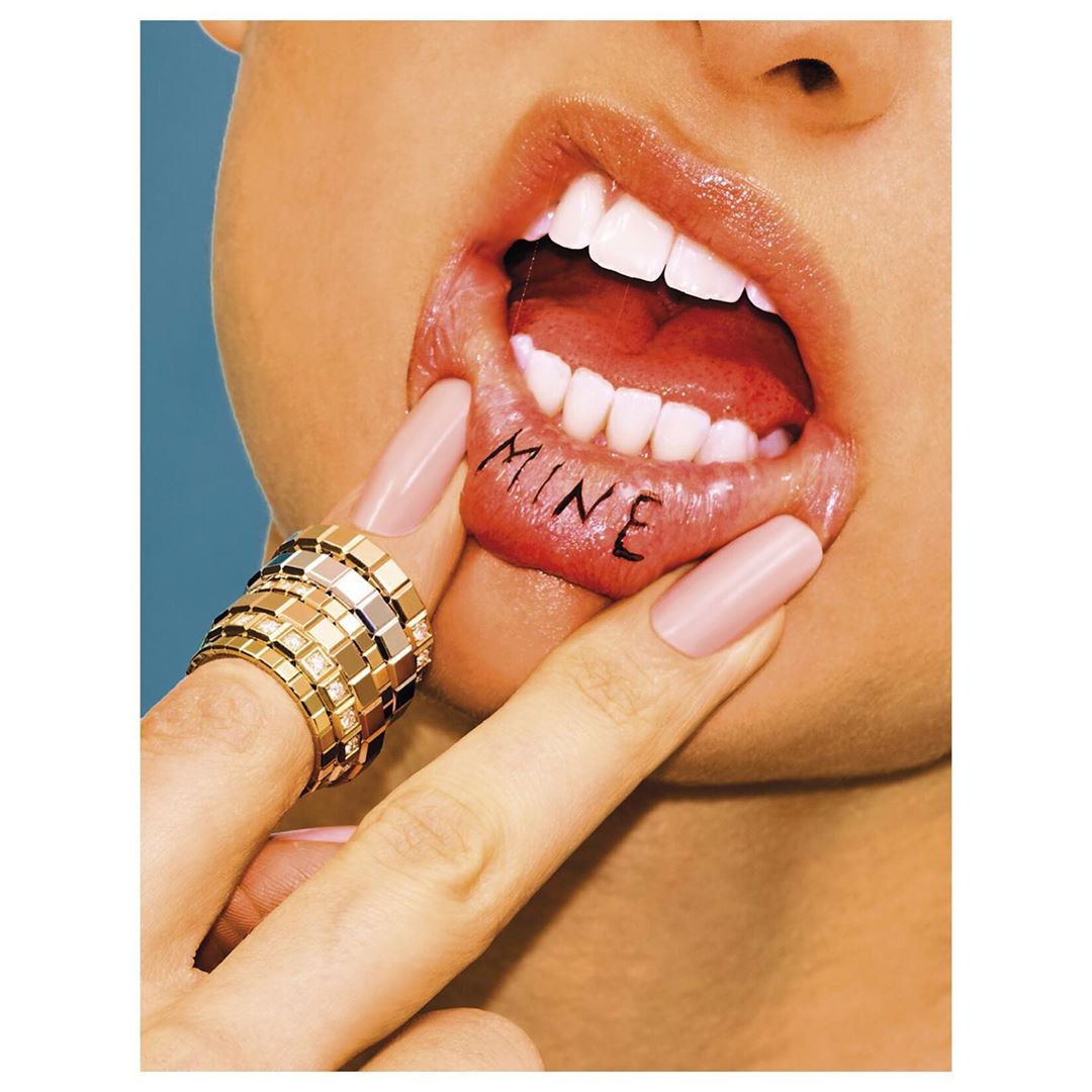 Close-up of a woman's face featuring open lips with a 'MINE' tattoo on her lower lip, a gold ring on her finger, and a blue background.