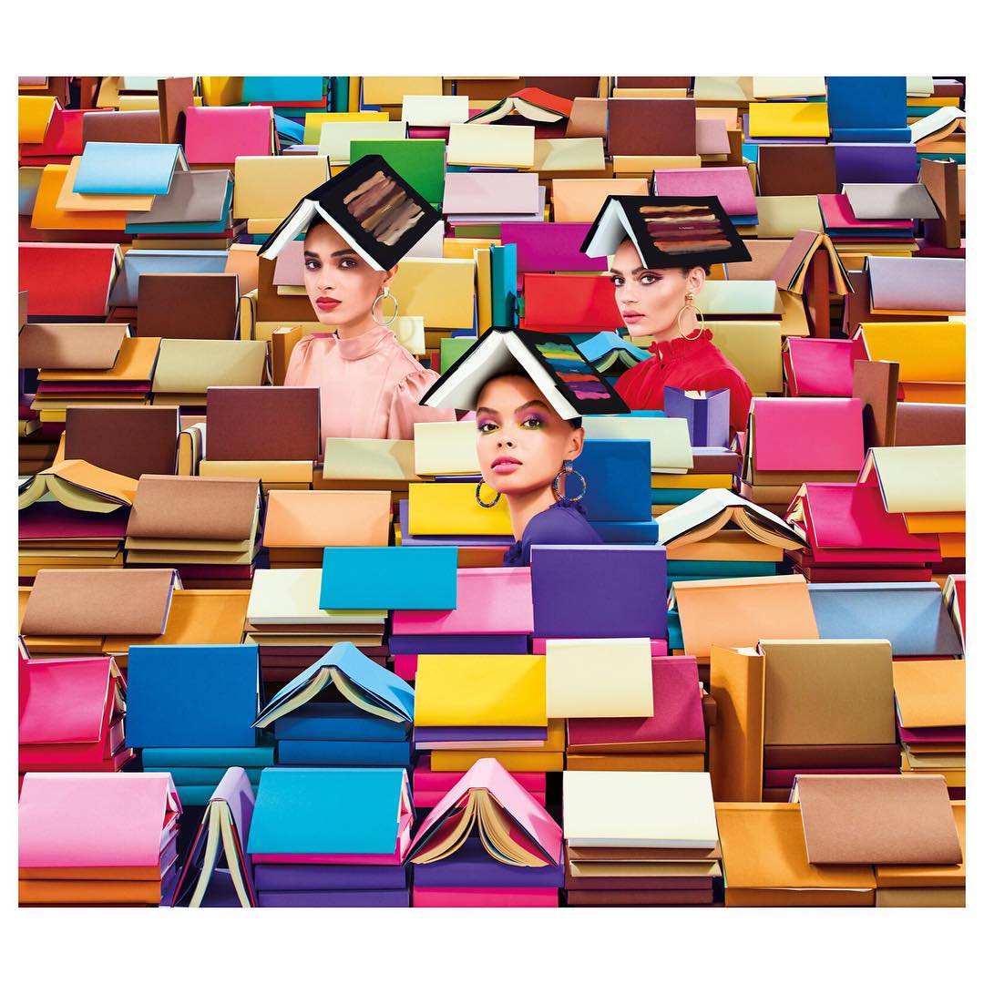 Three women pose amidst a sea of colorful books, some worn as hats, in a visually striking and imaginative editorial composition.