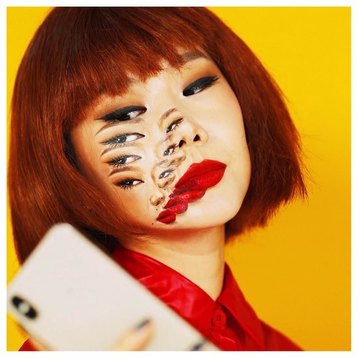 A young adult female with red hair displays intricate face art makeup featuring multiple eyes and red lips, holding a smartphone.