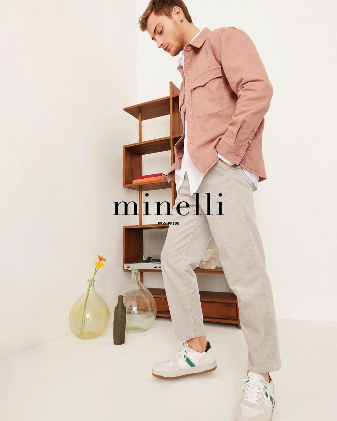 Young man in a pink jacket, light pants, and sneakers, standing in a neutral-toned room with a wooden shelf and glass vases.