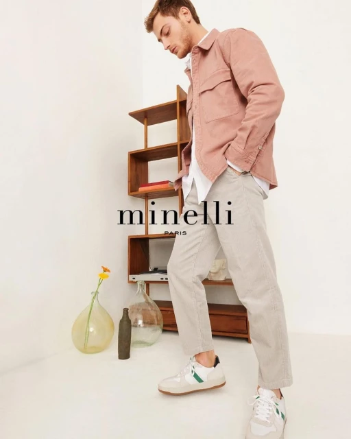 Young man in a pink jacket, light pants, and sneakers, standing in a neutral-toned room with a wooden shelf and glass vases.