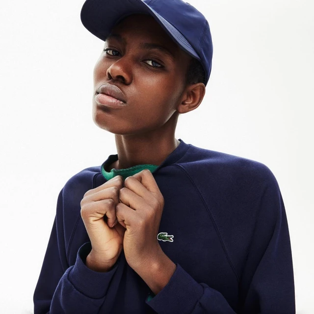 Headshot of adult woman in blue cap and navy sweatshirt with Lacoste logo, posing against a white background in a fashion setting.