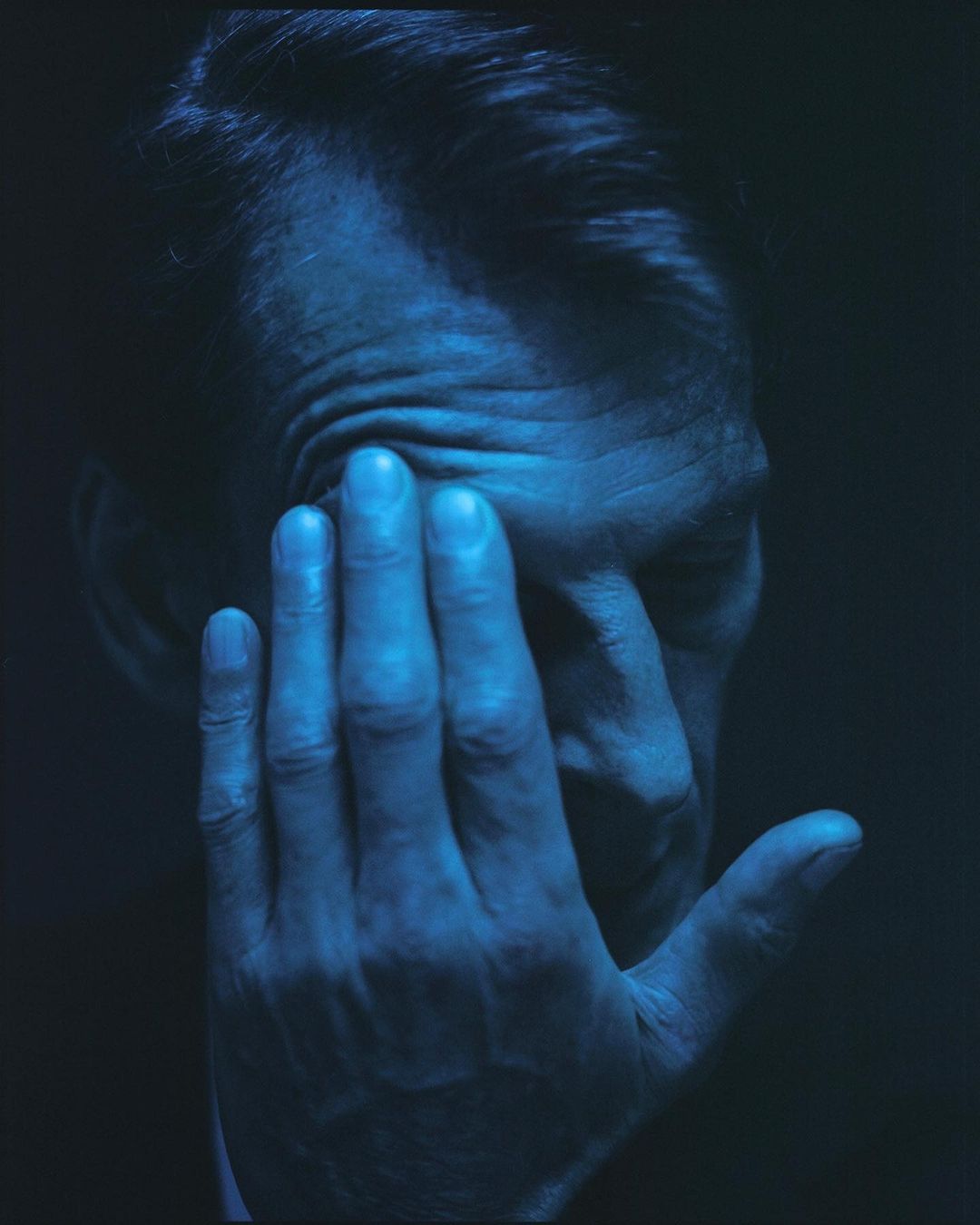 Dramatic portrait of an adult male with hand to face, shot in deep blue hues and dark lighting, highlighting skin texture and form.