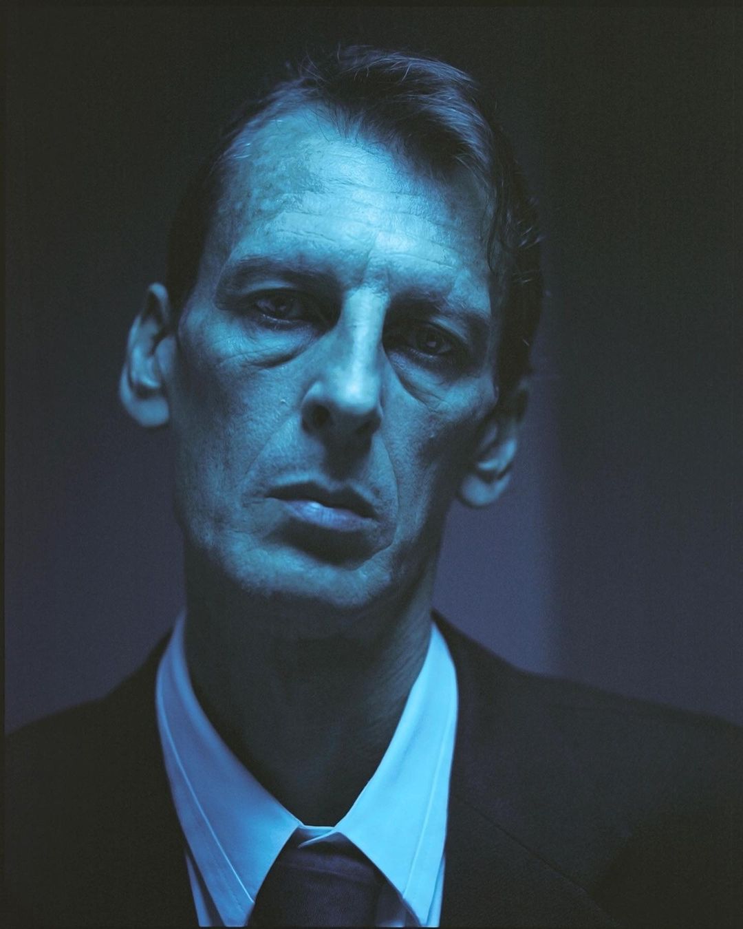 Headshot of a serious-looking man with short brown hair wearing a dark suit, white shirt, and dark tie under blue-toned lighting.