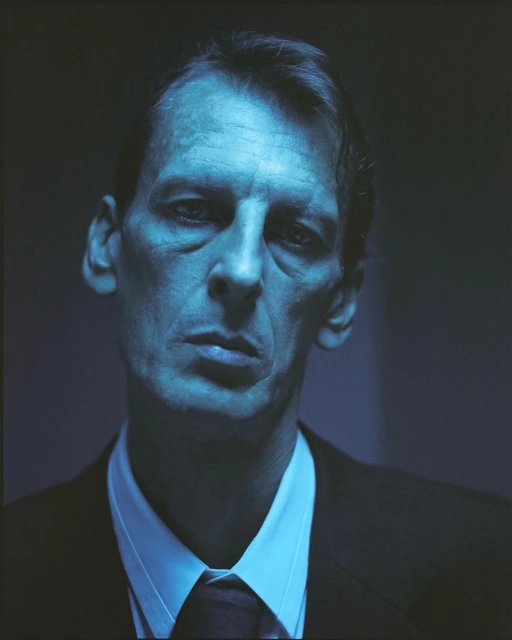 Headshot of a serious-looking man with short brown hair wearing a dark suit, white shirt, and dark tie under blue-toned lighting.