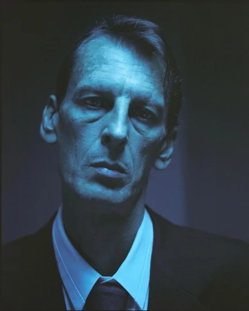 Headshot of a serious-looking man with short brown hair wearing a dark suit, white shirt, and dark tie under blue-toned lighting.