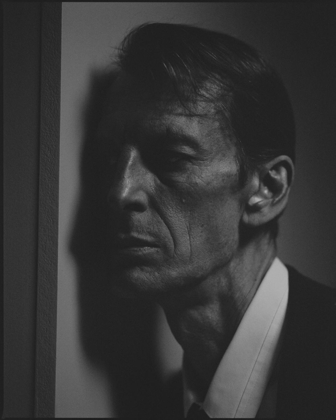 Monochrome portrait of a mature adult man in a suit and collared shirt with a dark background, shadowed face and side lighting.