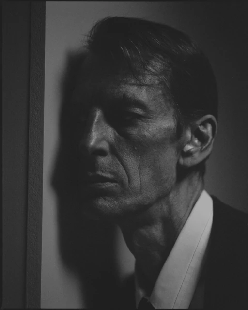 Monochrome portrait of a mature adult man in a suit and collared shirt with a dark background, shadowed face and side lighting.