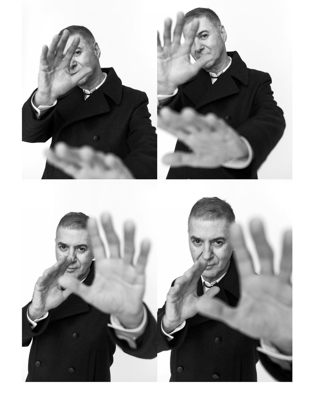 A black and white studio portrait of a mature adult man in a dark overcoat, posing with his hands up toward the camera.