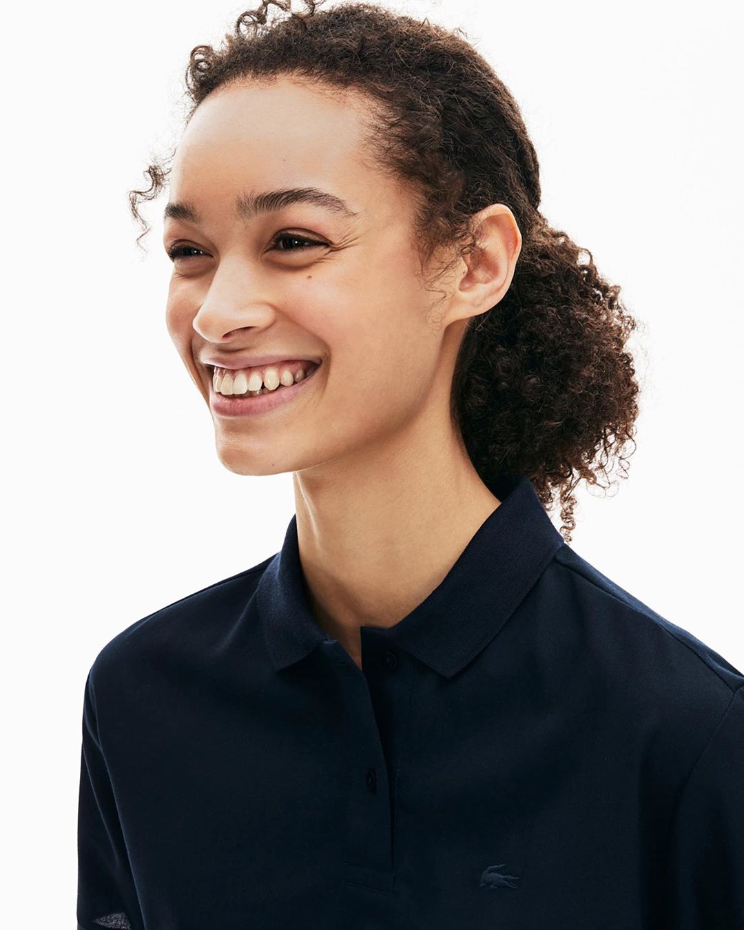 Close-up of a smiling woman with dark, curly hair wearing a dark blue collared long sleeve shirt against a white background.