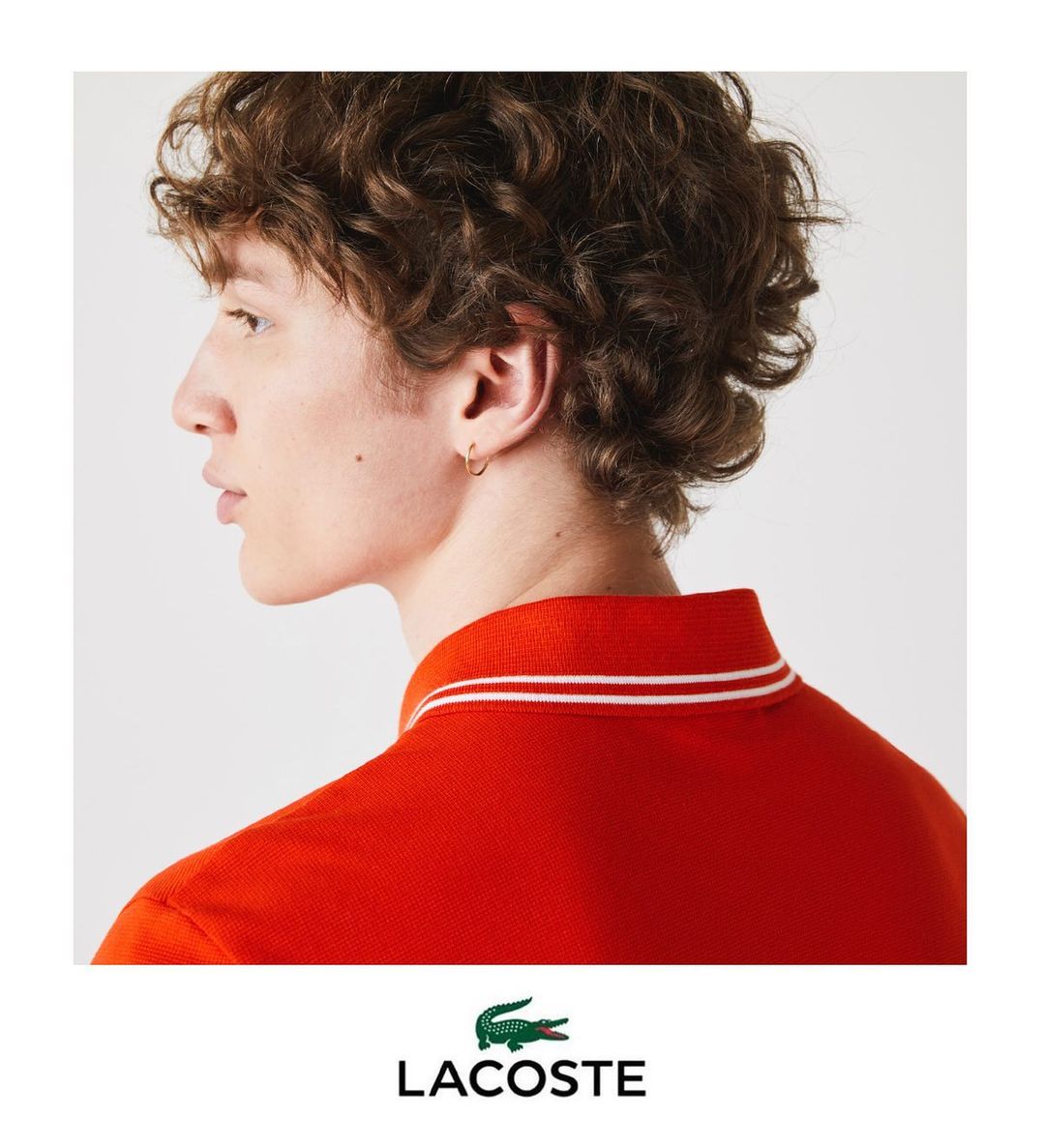 Profile of a curly-haired male model in a red polo shirt with white accents, isolated on a clean, bright white background. Fashion shot.
