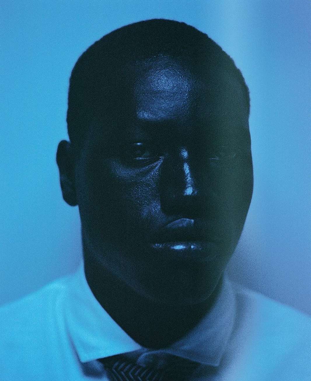 Portrait of a dark-skinned adult male wearing a white shirt and patterned tie, with a blue gradient background in soft studio lighting.