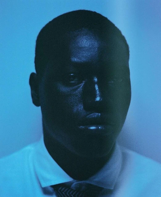 Portrait of a dark-skinned adult male wearing a white shirt and patterned tie, with a blue gradient background in soft studio lighting.
