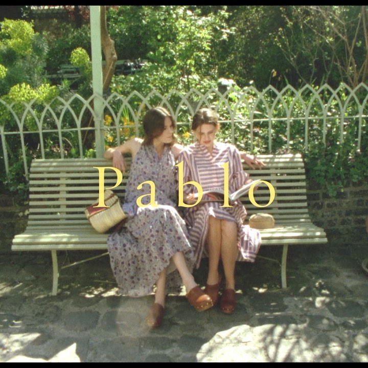 Two young women sit together on a park bench, wearing dresses and clogs, under dappled sunlight and the text overlay 'Pablo'.