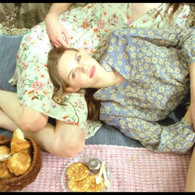 Two women in floral dresses share a picnic with pancakes and croissants, lying on a blue and pink blanket on a sunny day.