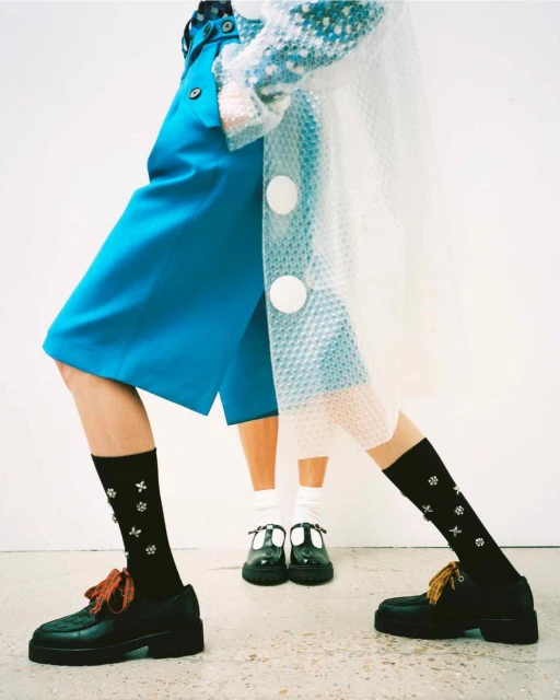 Fashion shot featuring two models wearing blue culottes, black socks with white floral pattern, and black loafers on a light grey floor.