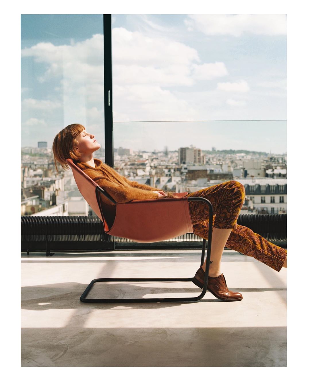 Adult woman relaxes in a chair on a balcony overlooking a European city skyline on a bright, sunny day with blue skies.