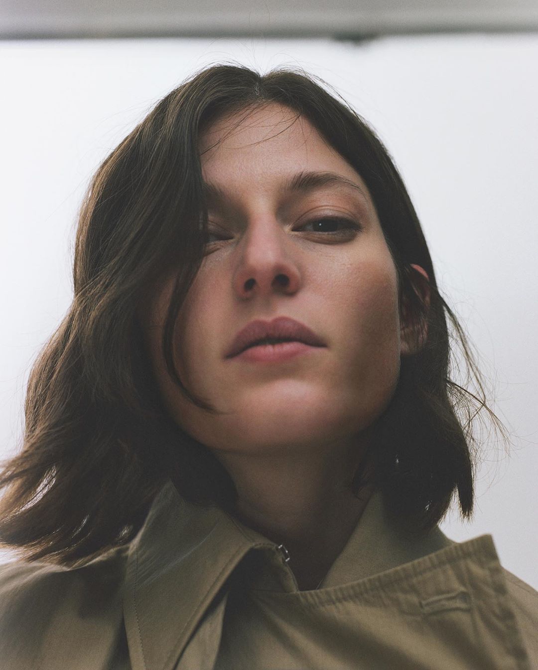 Close-up studio portrait of a woman with brunette wavy hair wearing a beige trench coat. She has fair skin and a neutral facial expression.