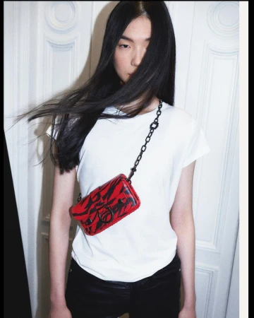 Woman with long dark hair wearing a white t-shirt and black jeans poses with a red and black patterned purse in front of a white door.