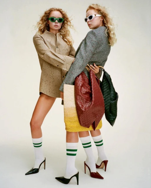 Two blonde women in sunglasses pose in fashionable attire with purses and heels in a studio setting against a neutral backdrop.
