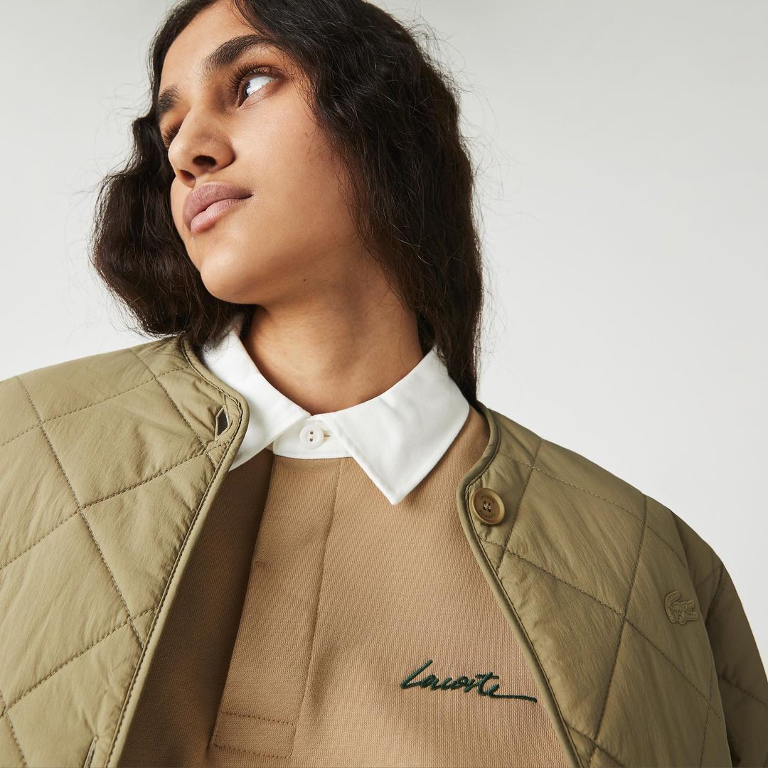 Close-up shot of a woman wearing a beige sweater with a white collared shirt underneath and a quilted, khaki jacket. Studio lit.