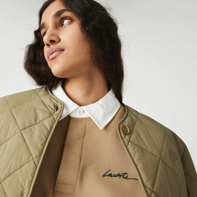 Close-up shot of a woman wearing a beige sweater with a white collared shirt underneath and a quilted, khaki jacket. Studio lit.