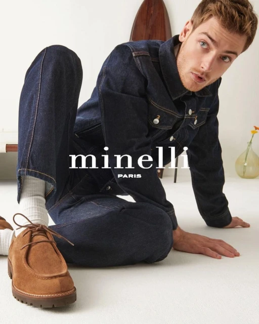 Man in denim jacket and jeans poses indoors next to brown suede shoes. Surfboard and clear glass vase in soft-lit background.