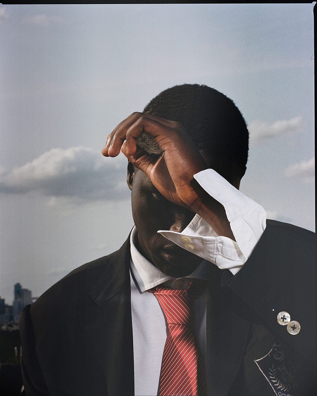 Portrait of an adult male wearing a black suit and red tie, shielding his eyes with his hand, with a cityscape in the background.