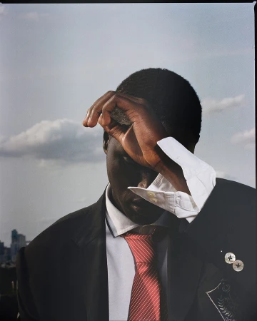 Portrait of an adult male wearing a black suit and red tie, shielding his eyes with his hand, with a cityscape in the background.