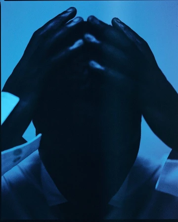 Person holding their head with hands in a dark blue image, wearing a white shirt with the collar visible against a blue background.