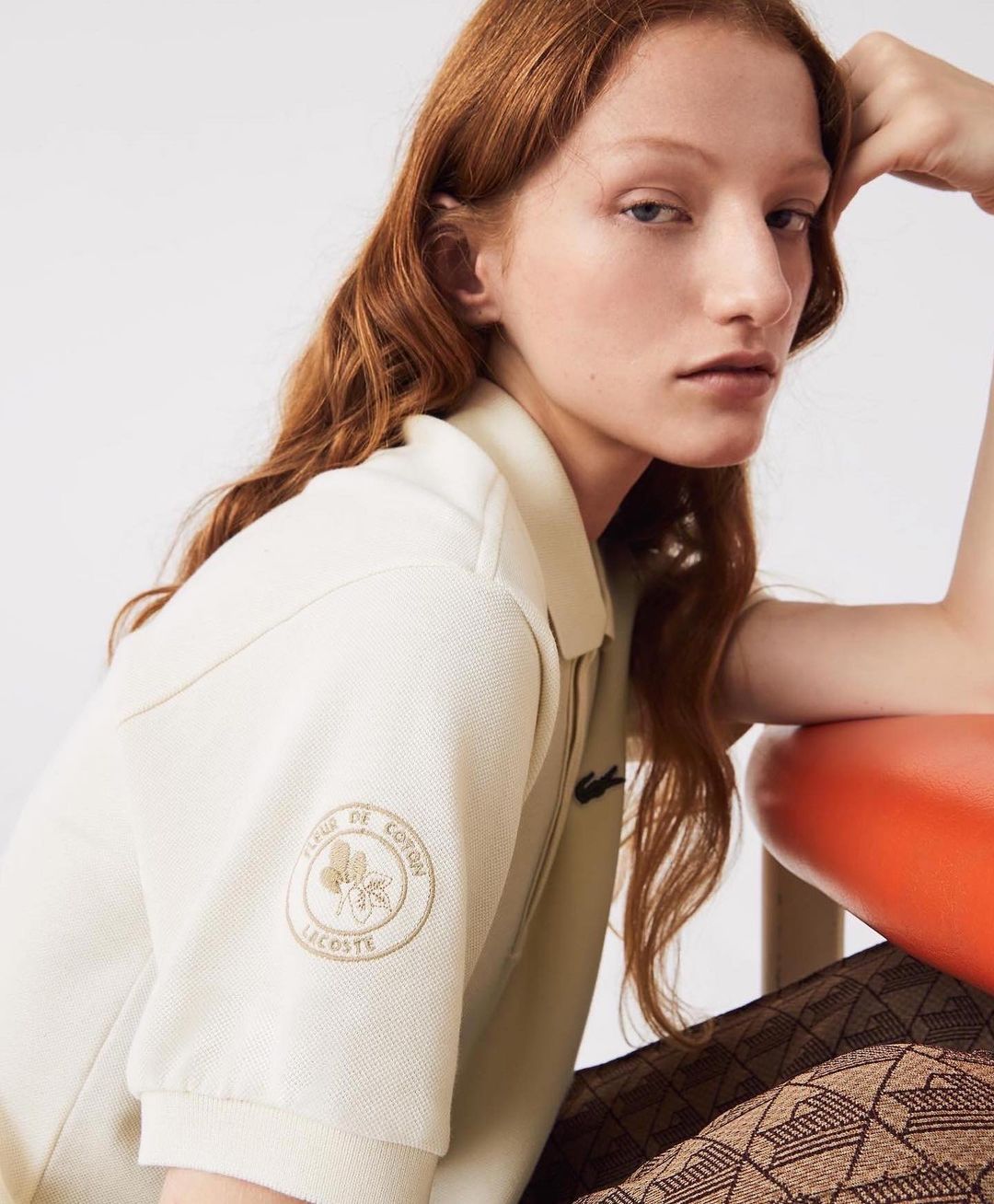 Woman with red hair poses indoors wearing a Lacoste polo shirt with a logo patch, seated on an orange chair with patterned tights.