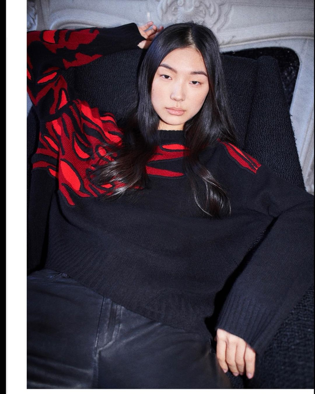 Portrait of a young Asian woman in a black sweater with red flame design, posing in an armchair against a textured white background.