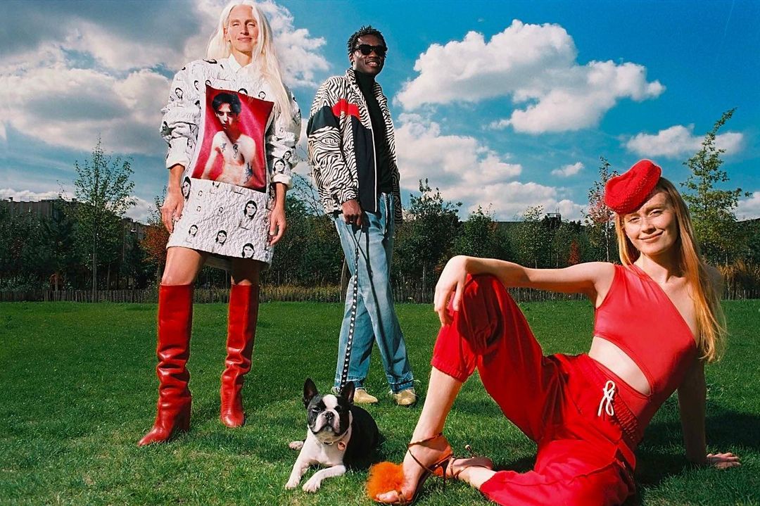 Three stylish people pose on a green lawn, with a Boston Terrier dog, under a cloudy blue sky in an outdoor setting.