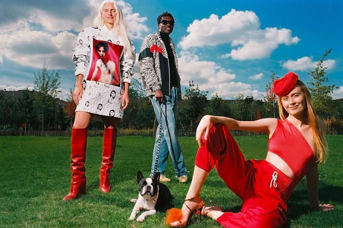 Three stylish people pose on a green lawn, with a Boston Terrier dog, under a cloudy blue sky in an outdoor setting.