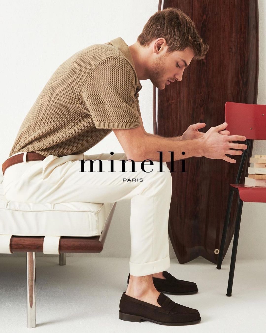 A man in a tan polo shirt and white pants sits on a bench wearing brown suede loafers in a fashion editorial-style shot.