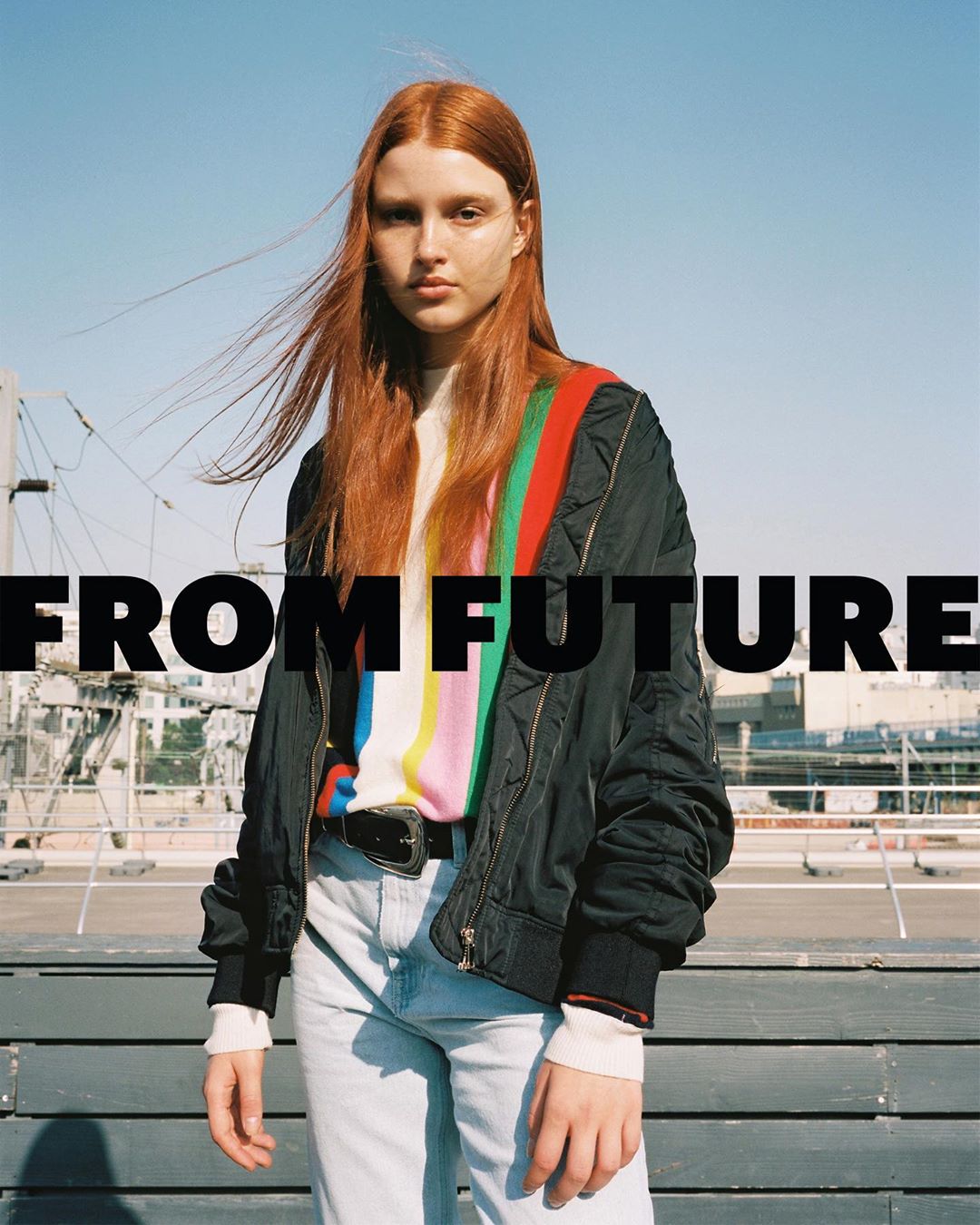 A waist-up shot showcases a young woman with red hair in a black bomber jacket and rainbow sweater against a clear blue sky.