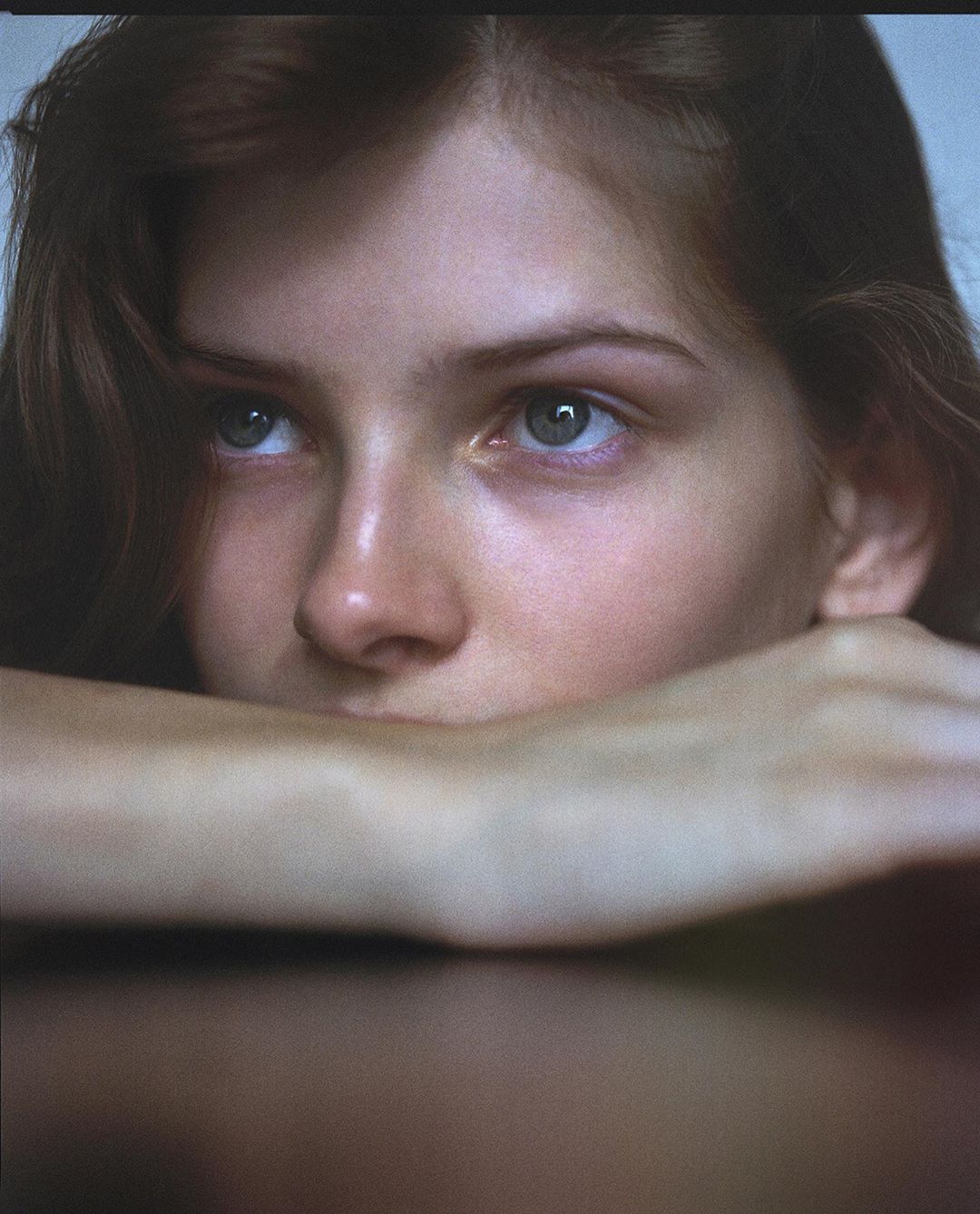 Close-up portrait of a young adult female resting her face on her arms with a serene expression, soft lighting, and muted color palette.