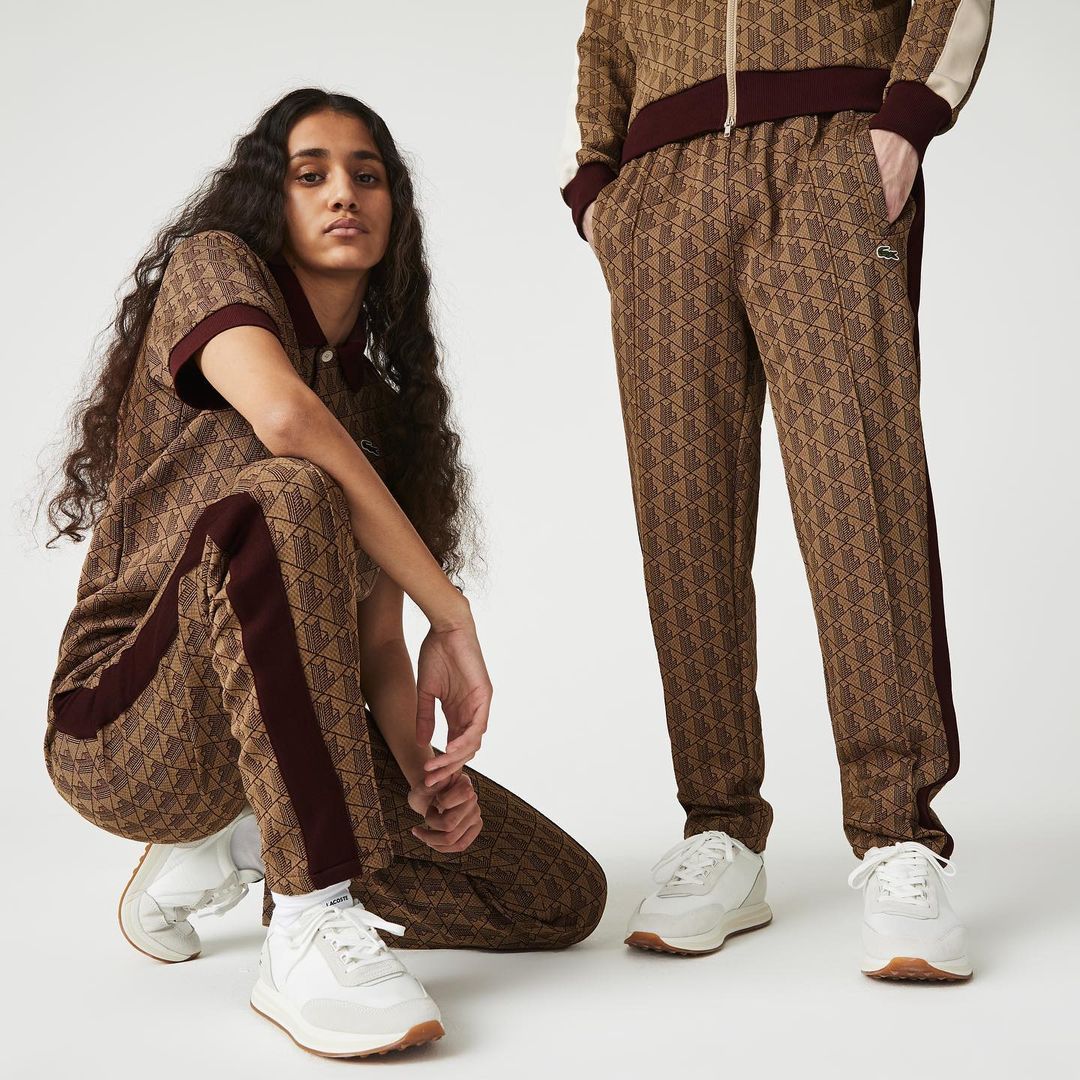 Two people wearing patterned Lacoste tracksuits and white sneakers in a studio setting, showcasing fashion apparel and sporty style.