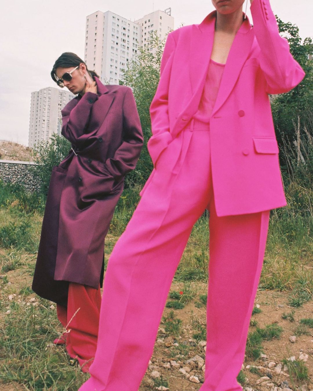 Two models outdoors in Paris, one in a vibrant pink suit and another in a plum coat standing before apartment blocks.