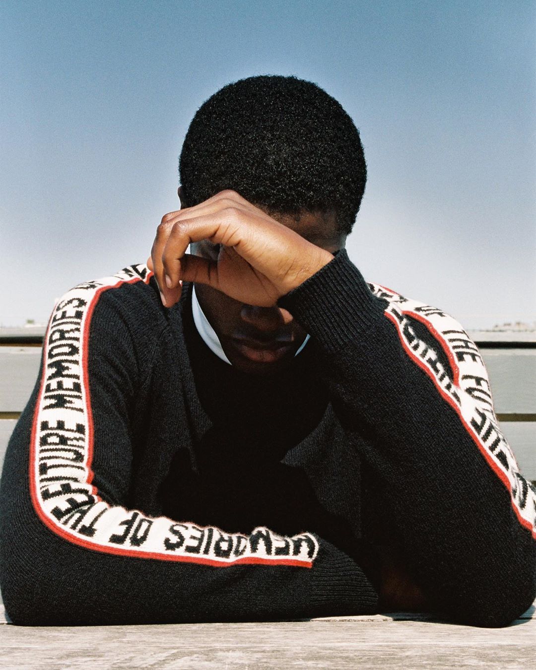 An African American man in a black sweater sits outdoors at a table, his hand obscuring part of his face. Clear sky background.